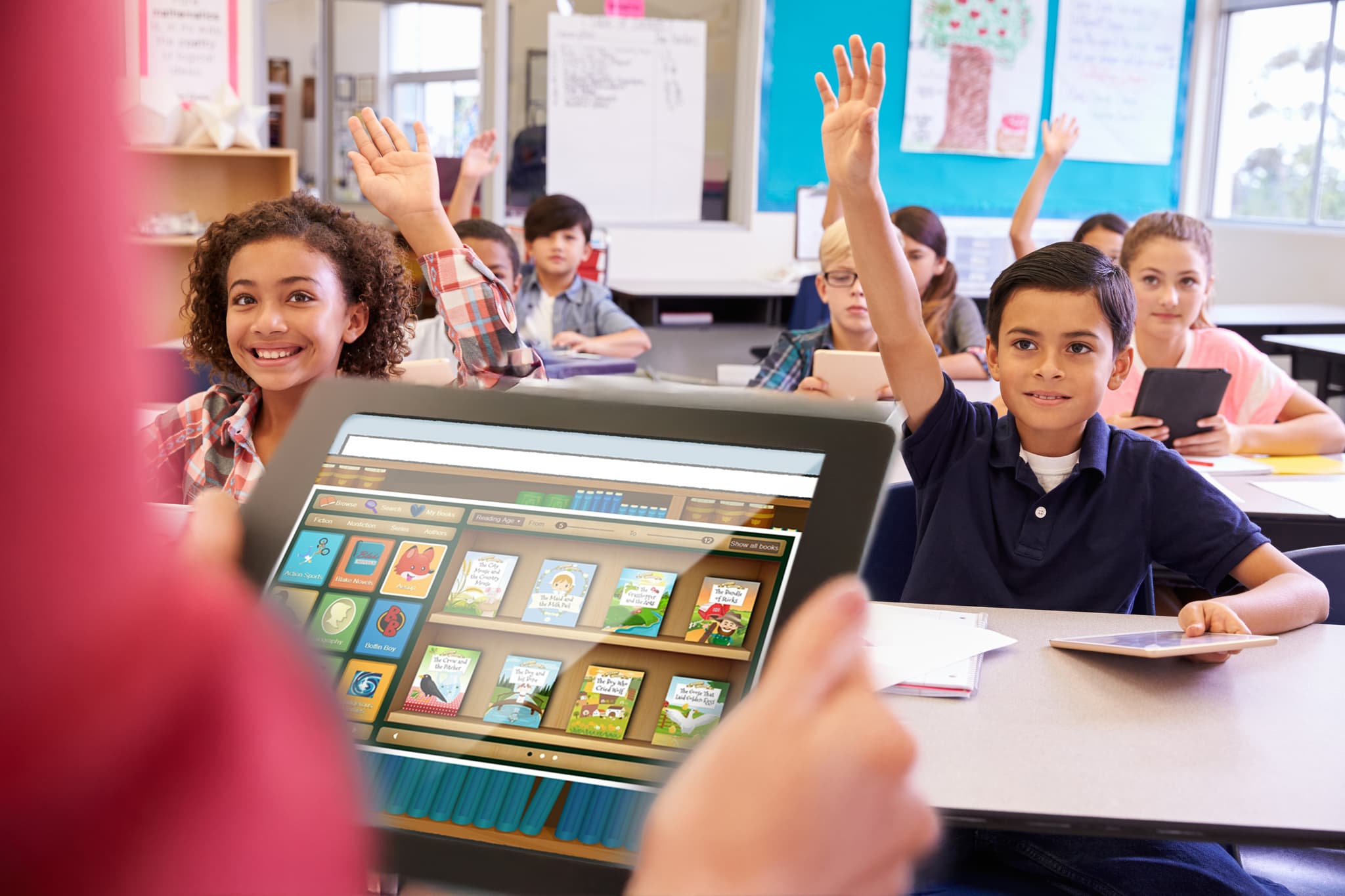 Primary school students raising their hands during a digital reading lesson using a tablet.